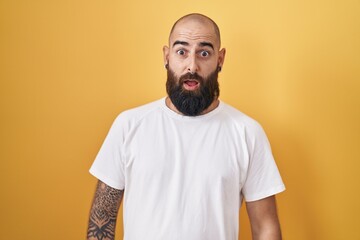 Young hispanic man with beard and tattoos standing over yellow background afraid and shocked with surprise and amazed expression, fear and excited face.