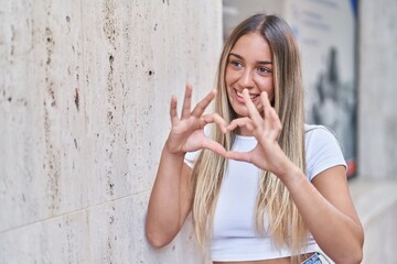 Young beautiful hispanic woman smiling confident doing heart gesture with hands at street