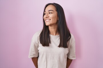 Young hispanic woman standing over pink background looking away to side with smile on face, natural...