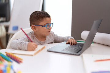 Adorable hispanic toddler student using laptop drawing on notebook at classroom