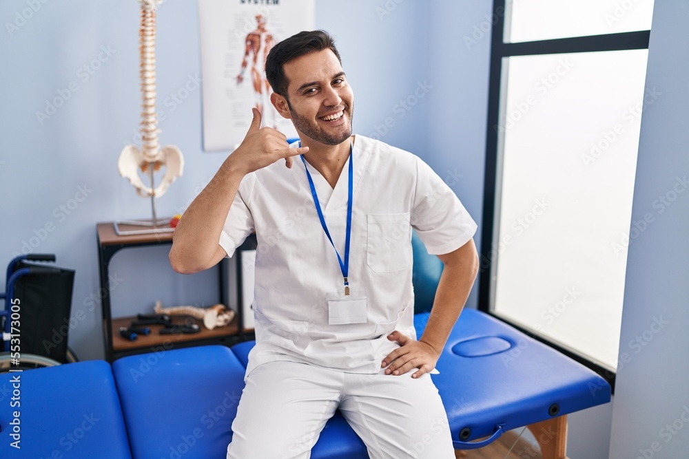 Wall mural young hispanic man with beard working at pain recovery clinic smiling doing phone gesture with hand 