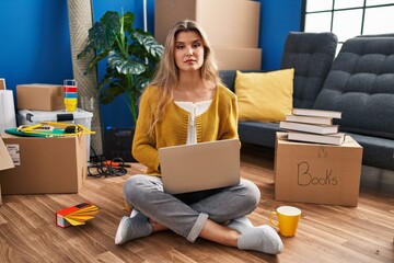 Young woman sitting on the floor at new home using laptop relaxed with serious expression on face. simple and natural looking at the camera.