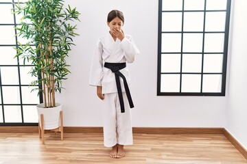 Young hispanic girl wearing karate kimono and black belt bored yawning tired covering mouth with...