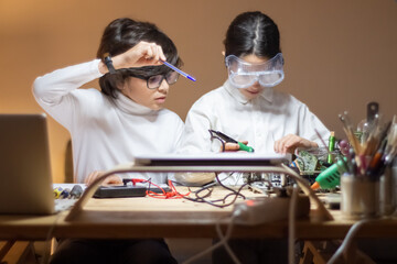 Serious girl in protective glasses using solder to create new electronic device. Boy giving advice...