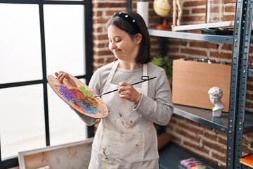 Young woman with down syndrome smiling confident holding paintbrush and palette at art studio