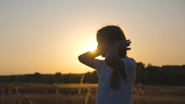 Happy small child look into camera standing in meadow and playing with her hair. Portrait of little smiling girl in grass field over sunset background. Concept of carefree and freedom. Dolly shot