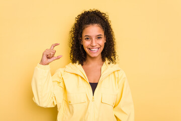 Young Brazilian curly hair cute woman isolated on yellow background holding something little with forefingers, smiling and confident.