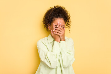 Young Brazilian curly hair cute woman isolated on yellow background blink at the camera through fingers, embarrassed covering face.