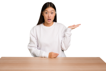 Young asian woman sitting on a table isolated impressed holding copy space on palm.