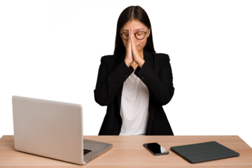 Young business asian woman sitting on a table isolated holding hands in pray near mouth, feels confident.