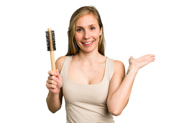 Young caucasian woman using an hair comb isolated showing a copy space on a palm and holding another hand on waist.