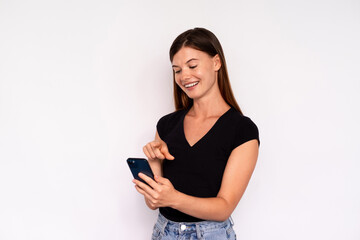 Naklejka premium Portrait of happy young woman surfing internet on smartphone over white background. Caucasian lady wearing black T-shirt and jeans texting message on mobile phone. Mobile communication concept