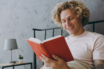 Young happy man wears white t-shirt pajama he sits in bed read novel book rest relax spend time in bedroom home in own room hotel wake up dream be lost in reverie good mood day. Real estate concept.