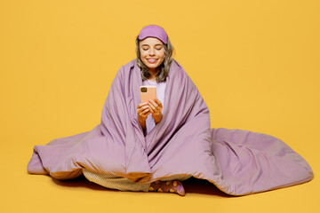 Fototapeta premium Full body young woman she wears purple pyjamas jam sleep eye mask rest relax at home sit wrap covered blanket duvet hold use mobile cell phone isolated on plain yellow background. Night nap concept.