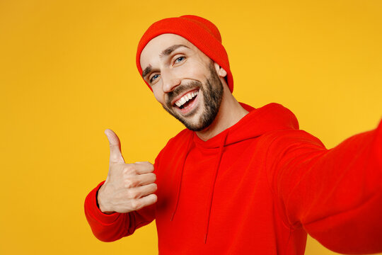 Close up young fun caucasian man wear red hoody hat doing selfie shot pov on mobile cell phone show thumb up gesture isolated on plain yellow color background studio portrait People lifestyle concept