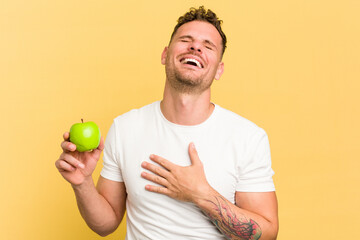 Young caucasian man eating an apple isolated laughs out loudly keeping hand on chest.
