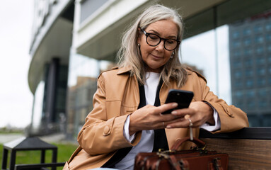 qafqazinfo senior Woman with a mobile phone in her hands on the background of the building sits on a bench