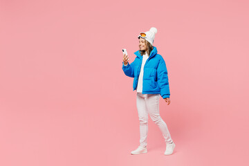Snowboarder woman wear blue suit goggles mask hat ski padded jacket walk go use hold mobile cell phone isolated on plain pastel pink background. Winter extreme sport hobby weekend trip relax concept. © ViDi Studio