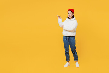 Full body happy smiling young woman of Asian ethnicity wear white padded windbreaker jacket red hat pointing indicate on workspace area copy space mock up isolated on plain yellow background studio.