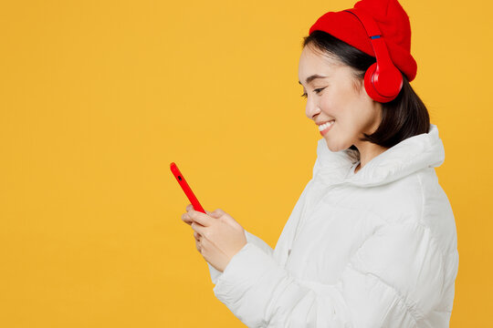 Side profile view young happy woman of Asian ethnicity wear white padded windbreaker jacket red hat headphones listen to music dance use mobile cell phone isolated on plain yellow background studio.