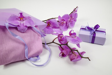 A gift in a lilac package and a decorative bag with a lilac shawl in it lie on a white table next to a blooming orchid
