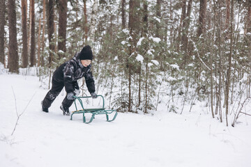 Happy teenager boy sledding and having fun outdoors. Joyful child playing in snow in winter forest. Laughing smiling kid walking in winter park in cold weather