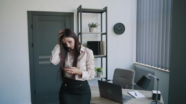 Young secretary worker businesswoman in shirt and skirt searching something on mobile phone in office. Shows different emotions