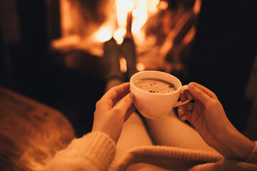 Woman drinkink hot cocoa sitting by the fire in front of cozy fireplace.
