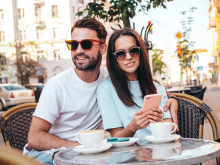 Smiling beautiful woman and her handsome boyfriend. Happy cheerful family. Couple drinking coffee in restaurant. They drinking tea at cafe in the street. Holding cup. Looking at smartphone, use apps