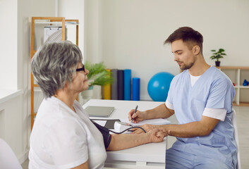 Obraz premium Young doctor measuring arterial blood pressure of senior patient. Elderly woman sitting at desk with tonometer cuff on her arm during visit to hospital. Health, medicine, disease prevention concept