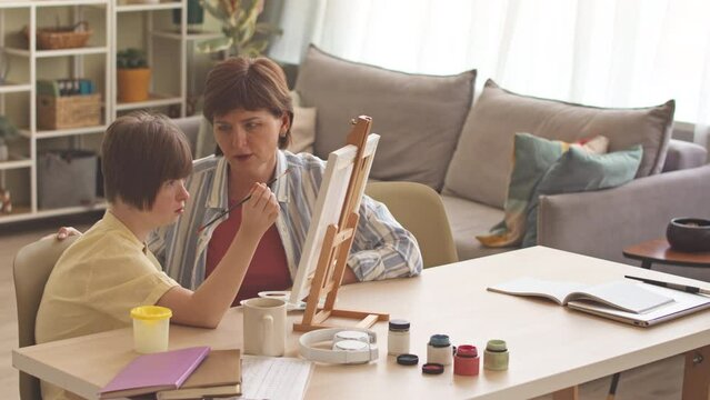Caring mother watching her daughter with down syndrome painting on canvas at home