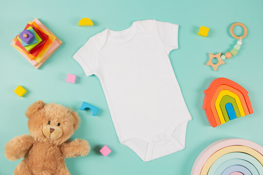 White cotton baby short sleeve bodysuit, teddy bear and natural wooden eco-friendly toys on green background. Infant onesie mockup. Blank gender neutral newborn bodysuit template mock up. Top view