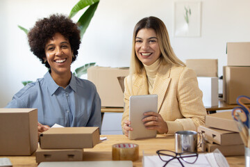 Obraz premium Portrait of logistics workers in office. Smiling young multi-ethnic colleagues sitting at table and examining parcels using tablet. Warehouse staff concept