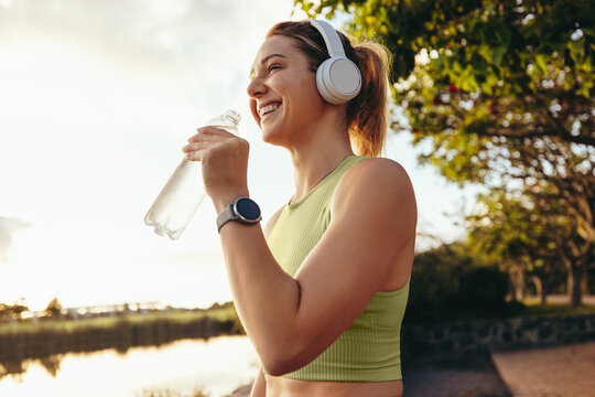 Female athlete taking a break outdoors, drinking water and listening to music with headphones