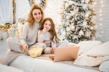 Young woman with child baby girl 7 years old in sleepwear watching film and eating popcorn on the bed in Christmas decorated home