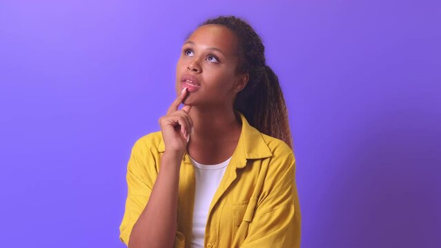 Young pensive casual African American woman zoomer dreamily touches chin and looks up coming up with plan for summer vacation or idea for shopping at mall stands on lilac studio background