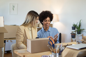 Multi-ethnic women processing orders online using digital tablet in home office. They sitting at table with cardboard box and discussing parcel tracking. Online shop concept