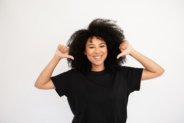 Cheerful young woman pointing at herself with thumbs. Caucasian female model with afro hairstyle and brown eyes in casual clothes smiling and pointing at herself. Self-assurance, confidence concept