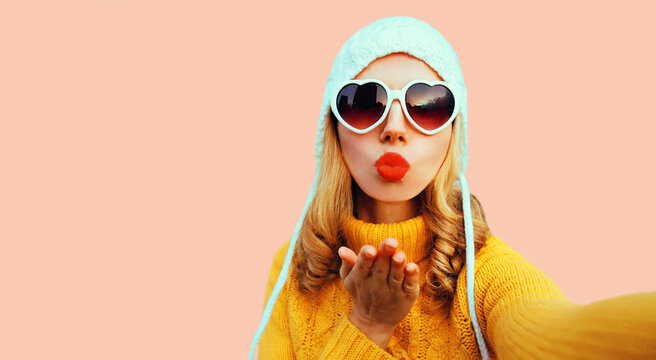Winter portrait of beautiful young woman stretching hand for taking selfie with smartphone wearing yellow knitted sweater, white hat, heart shaped sunglasses on pink background