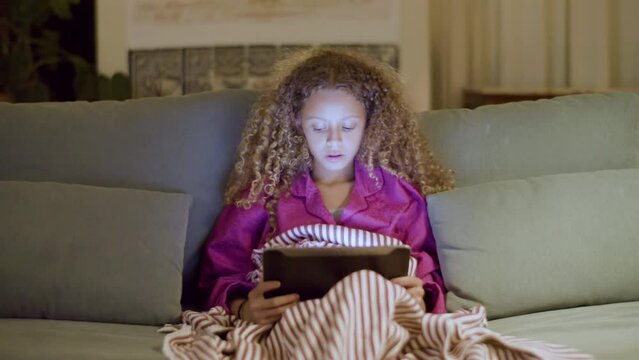 Young girl in pyjamas sitting on couch reading ebook. Pretty girl holding digital tablet on her knees in dimly lit room. Screen light reflecting on her face. Leisure, youth, modern technology concept.