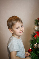 A photo of a beautiful boy in a gray T-shirt at the Christmas tree, looking into the camera. Portrait in a bright room. Natural, not staged photography.