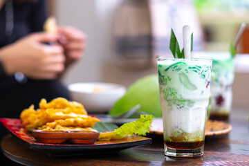 Es cendol is a traditional Indonesian iced dessert. Popular during fasting in Ramadan. A refreshing cold drink when drunk in the day and evening. A woman eating tempe as a bokeh background.