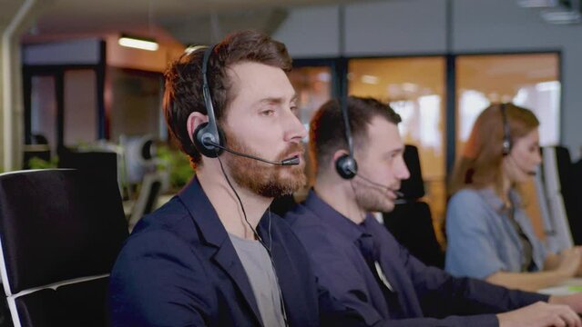 Close-up side view portrait of successful professional worker in call center talking with customers looking at camera smiling. Redhead bearded male operator using computer wearing headphones.
