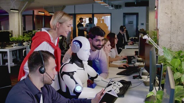 Side-view portrait of futuristic robot with AI working in office typing on keyboard using computer. IT workers smiling watching white scientific cyborg doing tasks. Artificial intelligence concept.