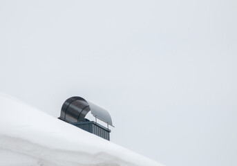 Smoke, metal exhaust pipe on the roof of a modern house. The roof of the building is covered with white snow against the gray sky. Cloudy, cold winter day, soft light.