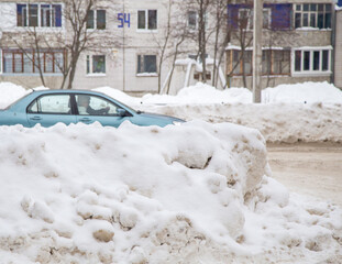 Large snowdrifts against the backdrop of a city street with a car. On the road lies white snow in high heaps. Urban winter landscape. Cloudy winter day, soft light.