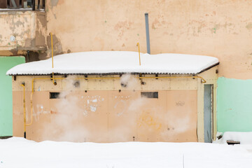 A round, metal exhaust pipe with smoke sticks out in the wall of the house. The roof of the building in white snow. Cloudy, cold winter day, soft light.