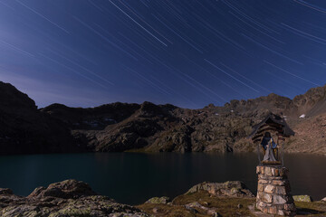 Star trails sky above alpine lake and statue