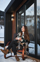 Young woman resting on terrace of modern barn house in the mountains. Happy female tourist sitting in chair, holding cup of tea, enjoying in new cottage in winter.