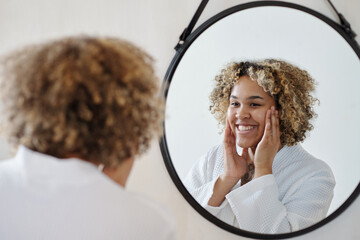 Young smiling black woman in white bathrobe touching her face while looking at her reflection in...